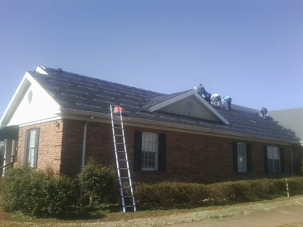 Crew tearing shingles off a brick ranch roof with a ladder set — Metal Roof Repair work by our crew in Lomax