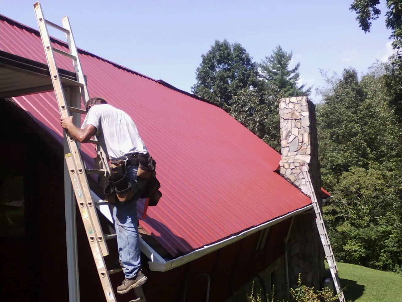 Roofer on a ladder beside a red metal roof and stone chimney — Soffit & Fascia Repair by Roofing Brothers in Lomax